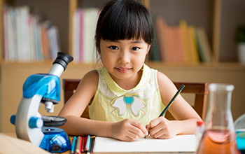 Girl writing on a notebook with a green pencil and a microscope next to her on a desk.